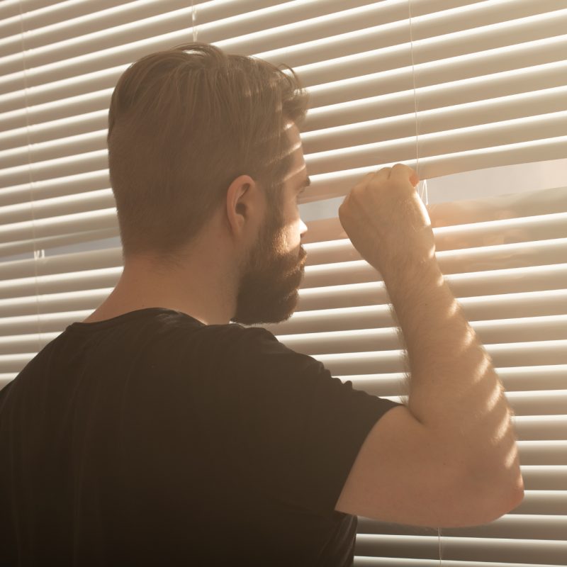 Rear view of young man with beard peeks through hole in the window blinds and looks out into the street. Surveillance and curiosity