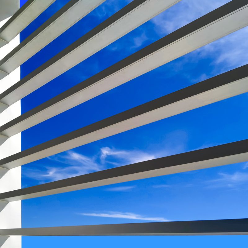 Sunlight on sunshade battens surface of office building against blue sky background, low angle view from inside of building