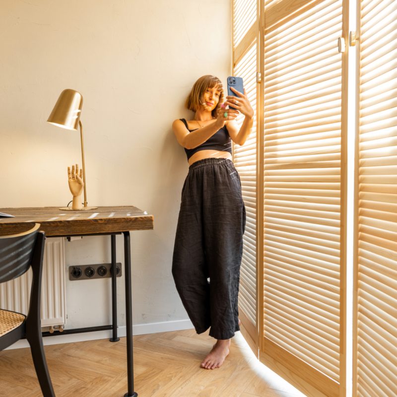 Young stylish woman in black home wear talking online or making photo on smart phone while standing near the window blinds and workplace at home. Concept of blogging and domestic lifestyle